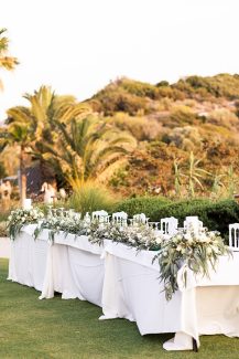 Πλούσιος στολισμός γαμήλιου τραπεζιού με πρασινάδες και λευκά ρομαντικά λουλούδια